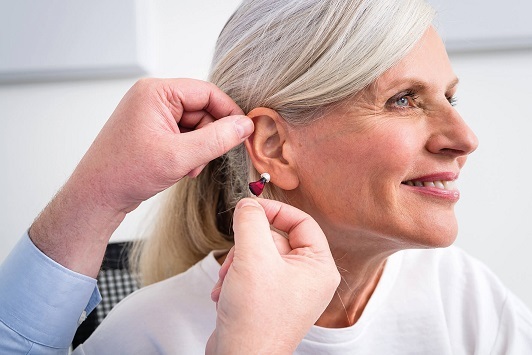 Ältere Frau bekommt von einem Hörakustiker ein Hörgerät eingesetzt und lächelt zufrieden.